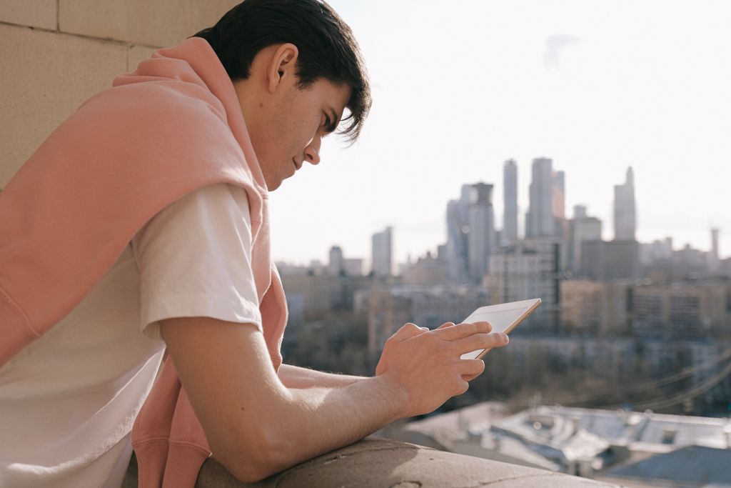 A young man stands on a balcony using a tablet with a city skyline in the background.