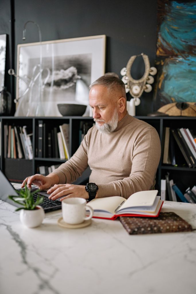 Mature man with beard working on laptop in a stylish home office with books and art.